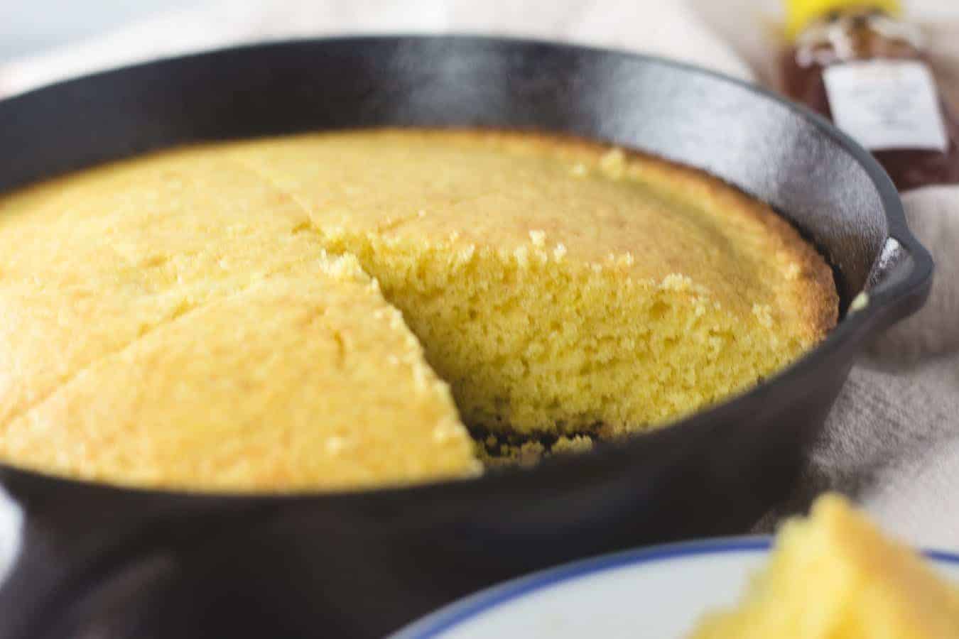 A close-up of cornbread in a black cast iron skillet with one slice removed. A jar of honey is blurred in the background.