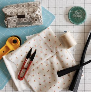 Sewing supplies on a grid cutting mat, including a rotary cutter, fabric scissors, spool of thread, needle case, measuring tape, patterned fabric, and a gray fabric pouch.
