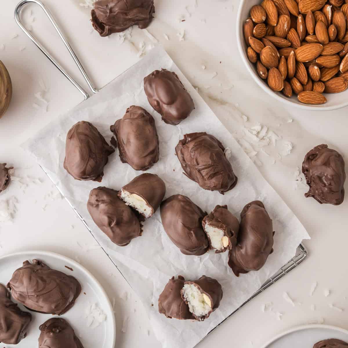almond joy bites on a wire rack, with some pieces broken open to show the white coconut filling, alongside a bowl of almonds.