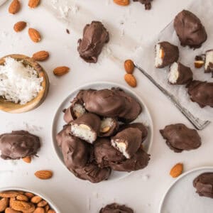 A pile of chocolate-covered coconut almond bites on a white plate, with some pieces cut open to reveal the interior, surrounded by loose almonds and a bowl of shredded coconut.