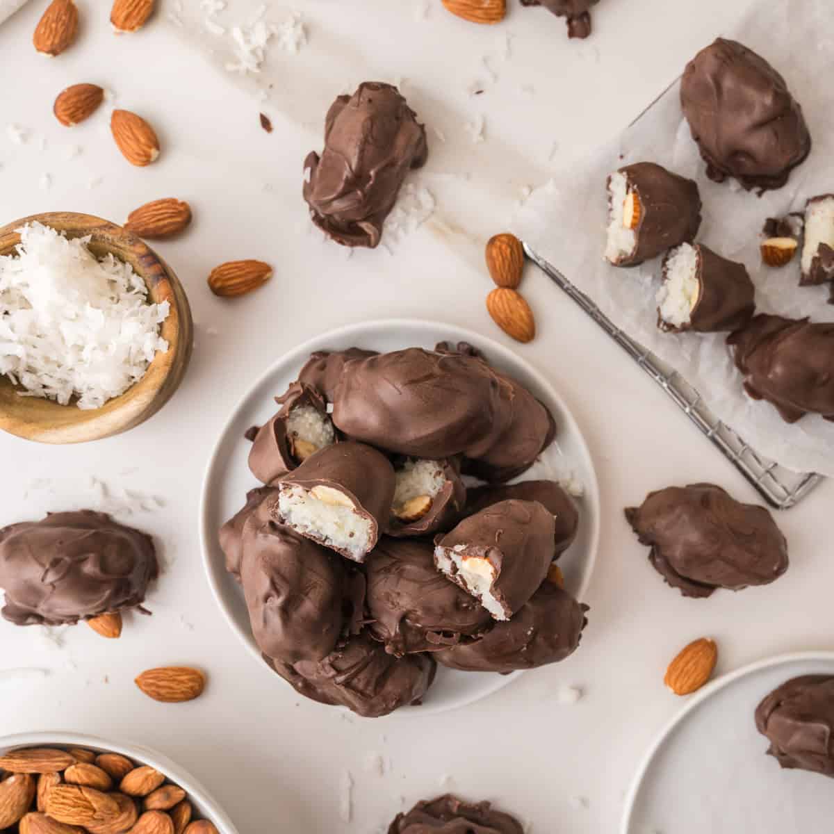 A pile of chocolate-covered coconut almond bites on a white plate, with some pieces cut open to reveal the interior, surrounded by loose almonds and a bowl of shredded coconut.