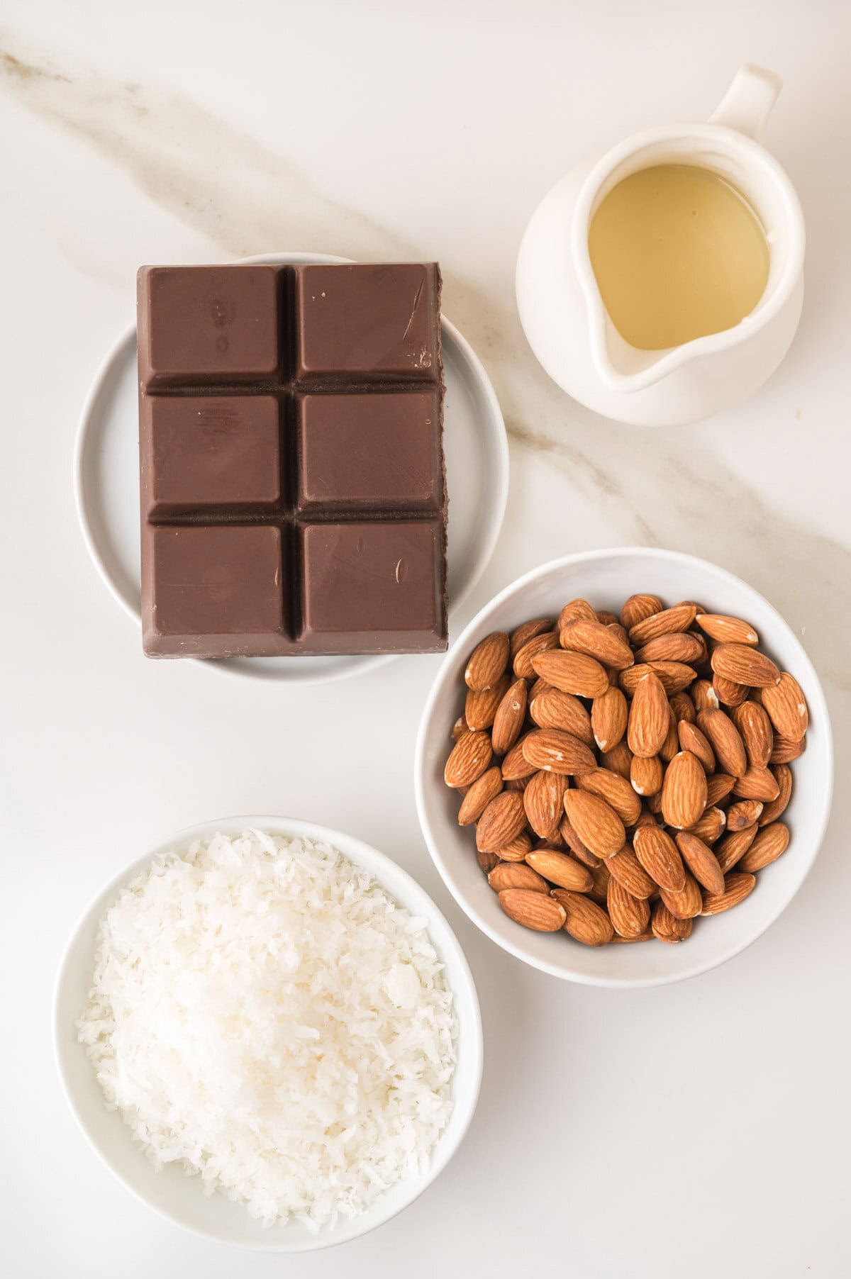 A plate with a chocolate bar, a small pitcher of sweetened condensed milk, and bowls of almonds and shredded coconut on a marble surface.