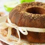 A bundt cake with a cinnamon sugar topping, wrapped with a cream-colored ribbon tied in a bow, sits on a plate.