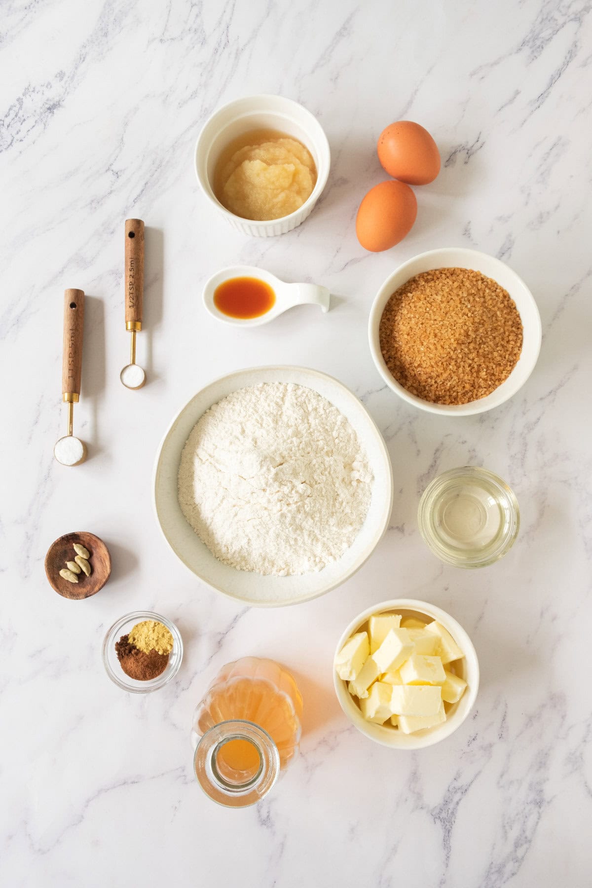 Baking ingredients on a marble surface: flour, eggs, butter, brown sugar, spices, apple sauce, oil, vanilla, and leavening agents, all measured in small bowls and cups, arranged neatly from above.