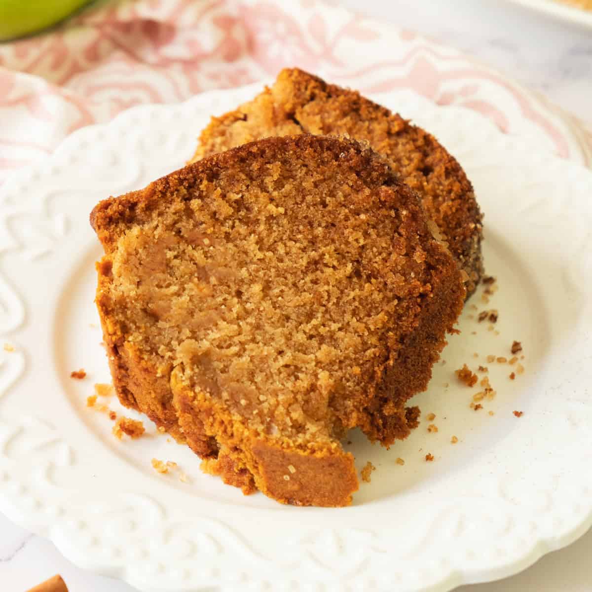 Two slices of moist, golden-brown apple bread sit on a white plate. Crumbs are scattered around, and a pink patterned cloth is partially visible in the background.