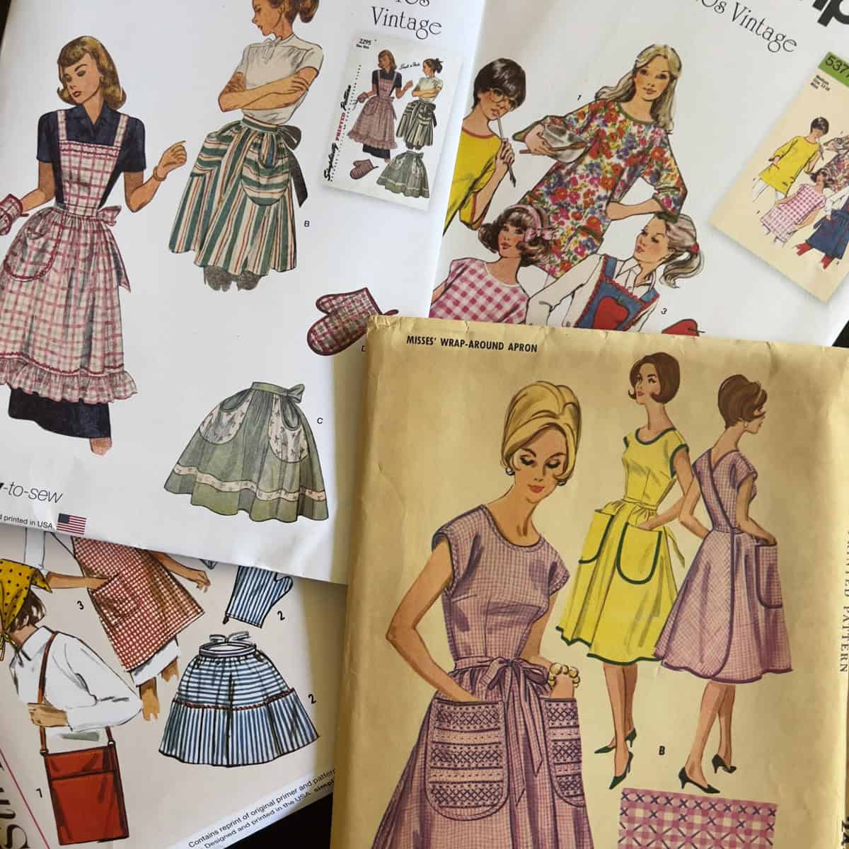 Assorted vintage apron sewing pattern envelopes and instructions featuring women in dresses and aprons, with some hand-drawn illustrations and colorful designs spread out on a surface.