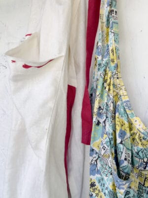 Two hanging aprons: a white apron with red trim on the left and a colorful, patterned fabric apron with houses and trees on the right, both against a light textured wall.