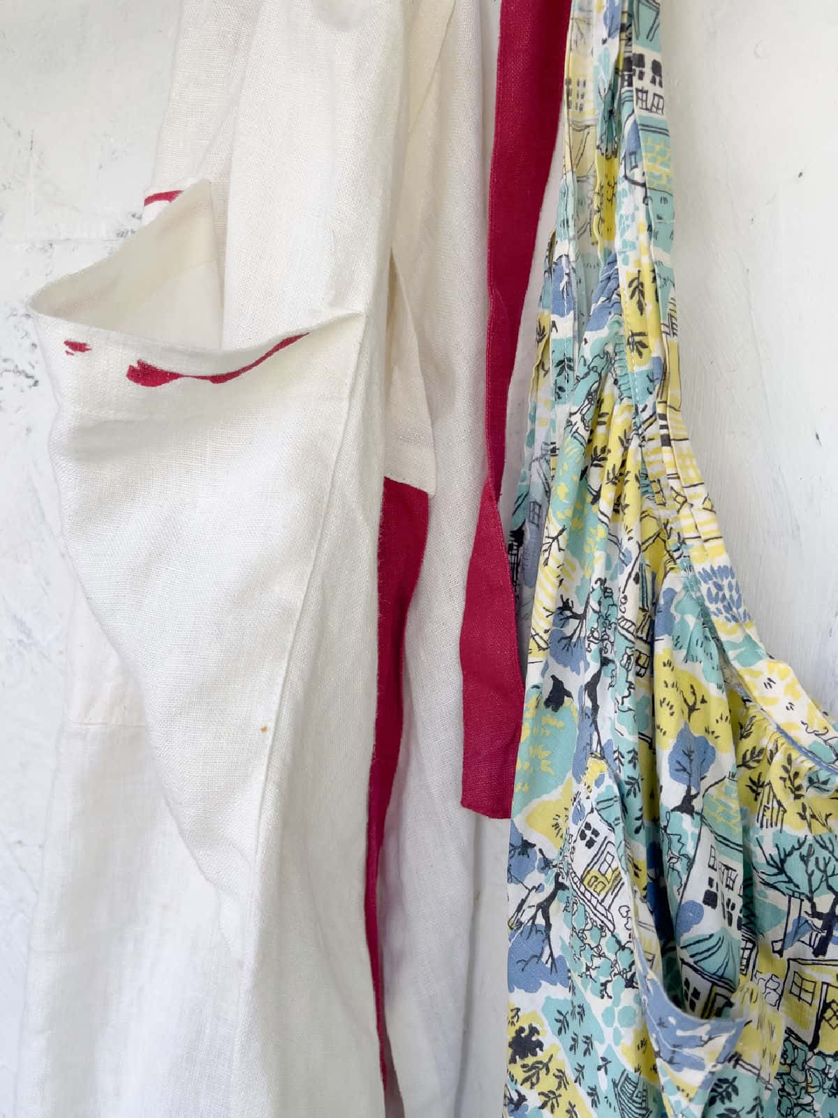Two hanging aprons: a white apron with red trim on the left and a colorful, patterned fabric apron with houses and trees on the right, both against a light textured wall.