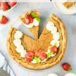 A Backwoods Maple Pie with a slice removed sits on a white surface, garnished with whipped cream and fresh strawberries. Two glasses of milk and scattered strawberries surround the pie.