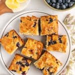 A plate with seven square pieces of blueberry cake, surrounded by a bowl of fresh blueberries, lemon slices on a board, and a floral napkin on a white surface.