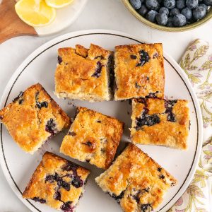 A plate with seven square pieces of blueberry cake, surrounded by a bowl of fresh blueberries, lemon slices on a board, and a floral napkin on a white surface.