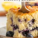 A fork cuts into a thick, golden blueberry biscuit on a plate, with fresh blueberries scattered around. The text above reads, “crumptiously delicious Blueberry Biscuits.”.