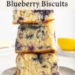 Three thick, homemade blueberry biscuits are stacked on a plate, showing a moist, fluffy texture with visible blueberries. The background is bright with a blurred yellow bowl and utensils. Text above describes Blueberry Biscuits.
