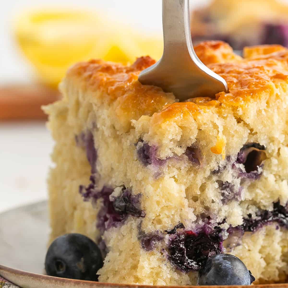 A close-up of a piece of moist blueberry cake on a plate, with a fork inserted into the top. Blueberries are visible in and around the cake, and a lemon is blurred in the background.