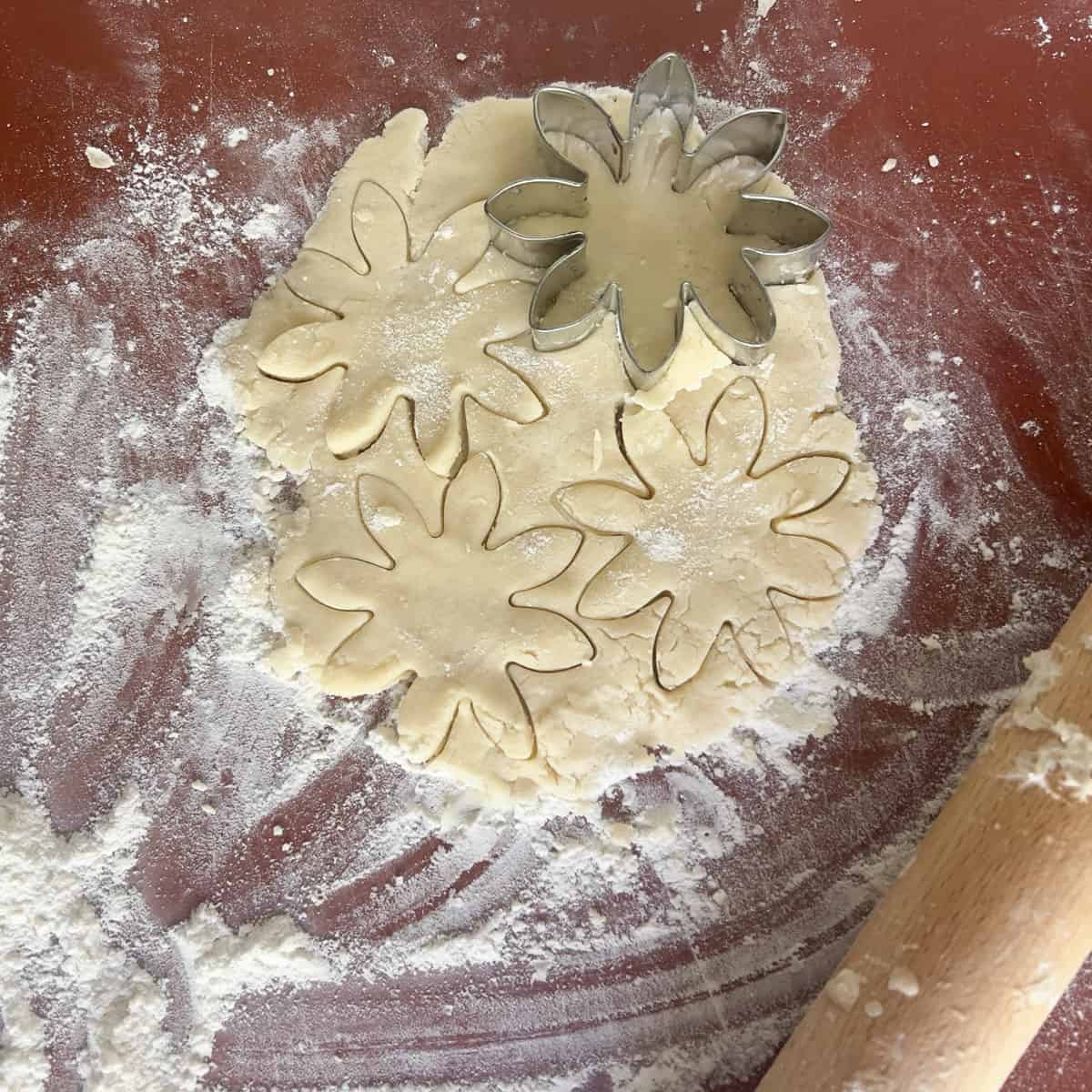 Cookie dough rolled out on a floured surface, with three flower shapes cut out and a metal flower-shaped cookie cutter resting on top. A wooden rolling pin is partially visible to the right.