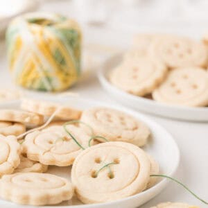 Plate of round sugar cookies designed to look like buttons, with some threaded with green string. A spool of yellow and green string and more cookies are visible in the background.