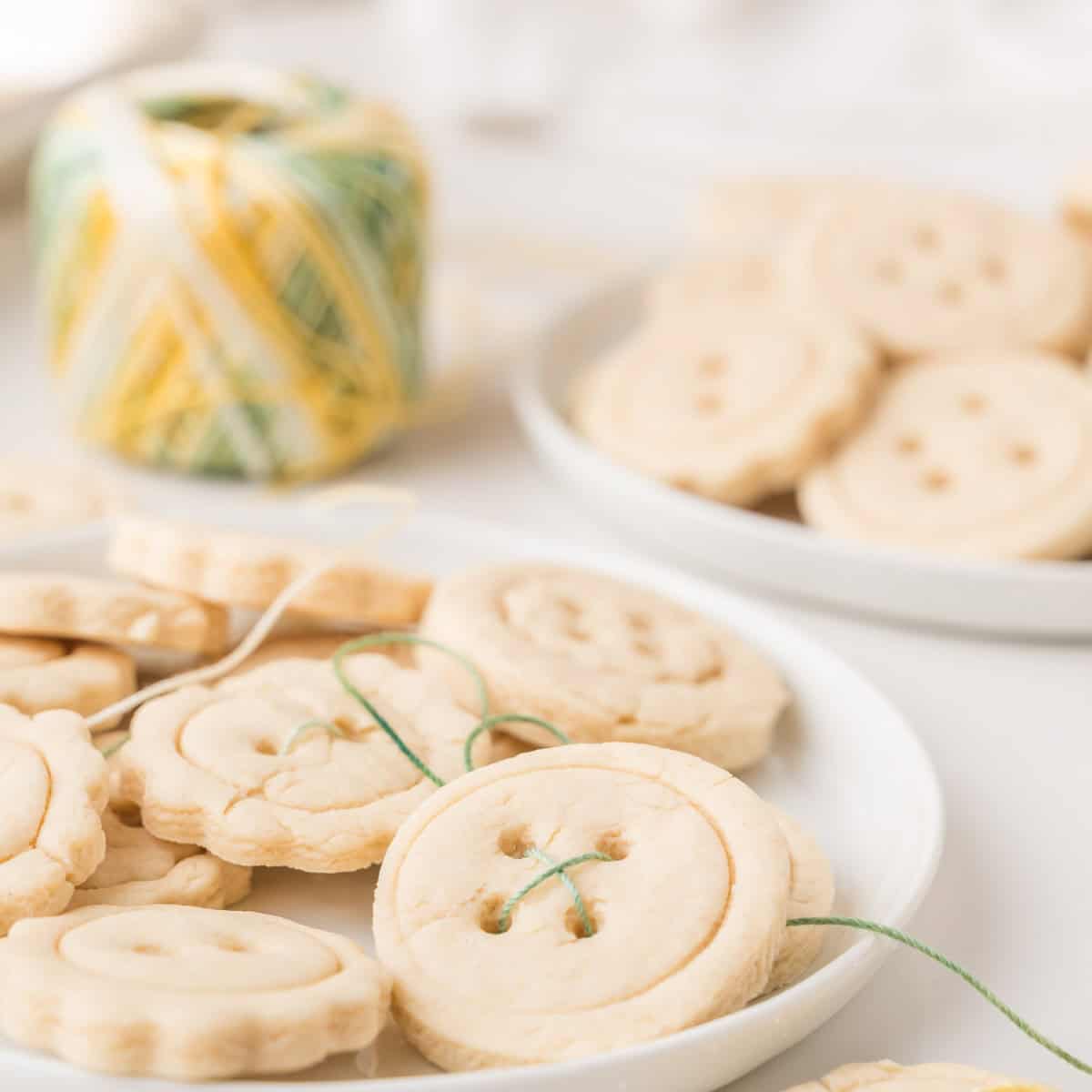 Plate of round sugar cookies designed to look like buttons, with some threaded with green string. A spool of yellow and green string and more cookies are visible in the background.