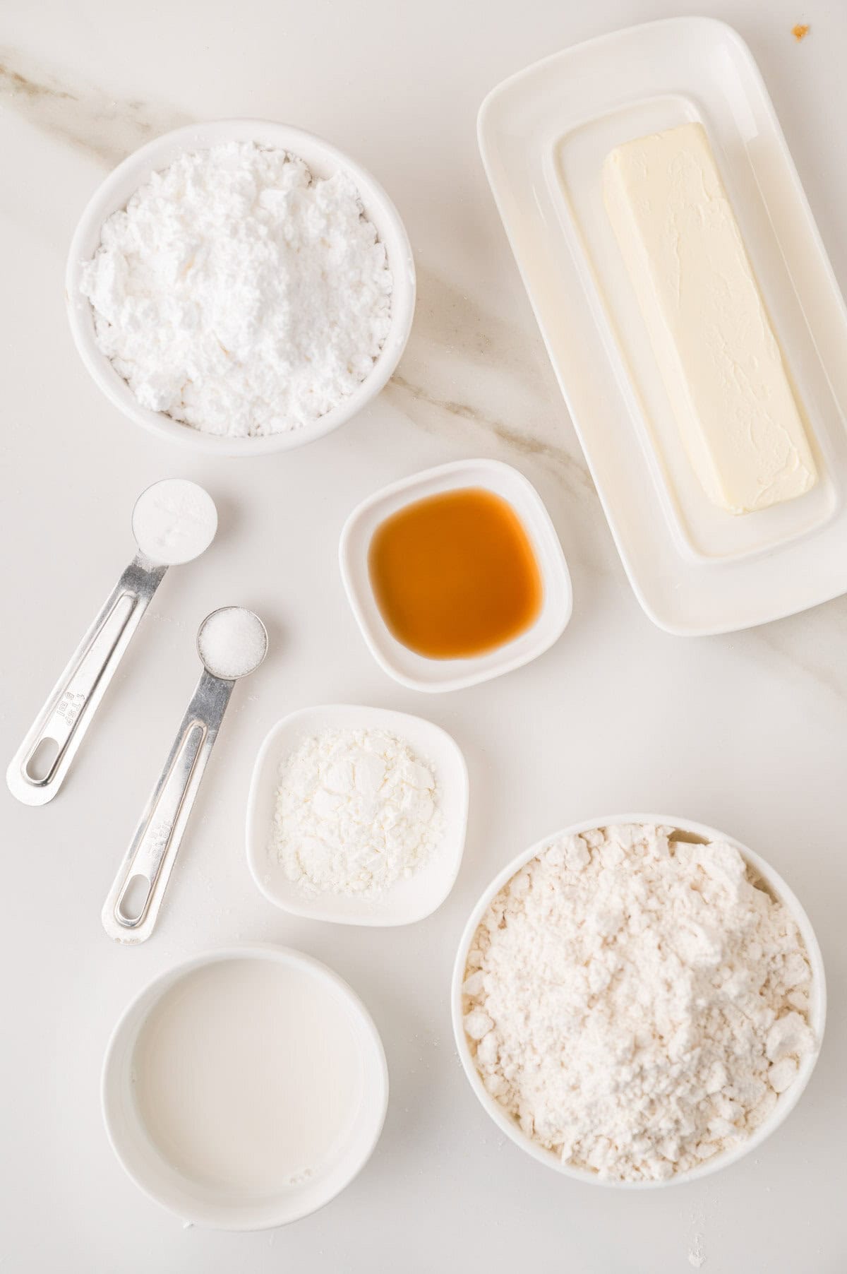 Small bowls and plates with flour, powdered sugar, butter, milk, vanilla extract, cornstarch, and two measuring spoons with salt and baking powder on a white surface.