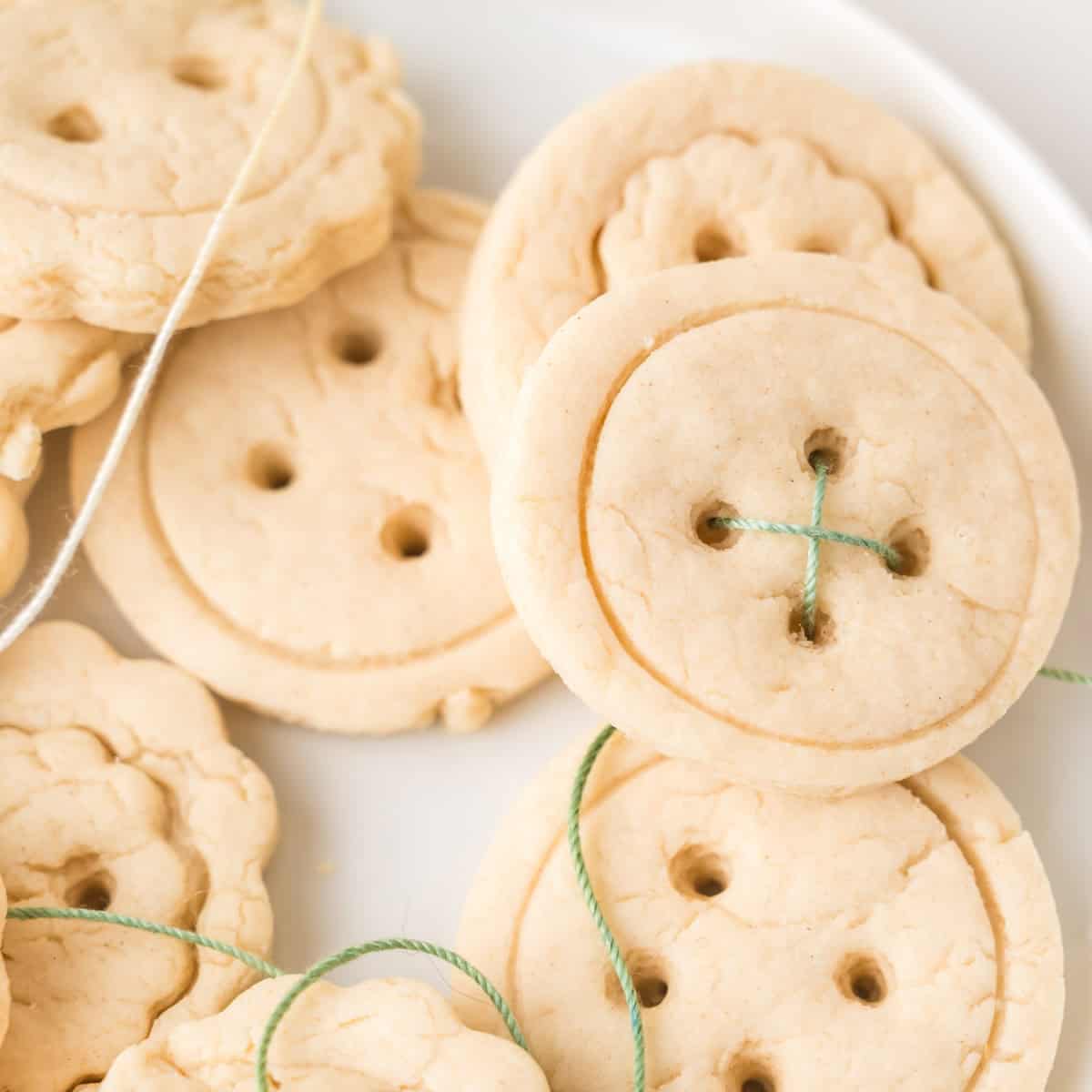Round sugar cookies designed to look like buttons, with holes in the center and green and white thread looped through some holes, displayed on a white plate.