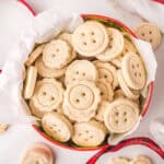 A round tin lined with white paper, filled with button-shaped sugar cookies, some with scalloped edges and holes to mimic button designs. A red ribbon is partially visible around the tin.
