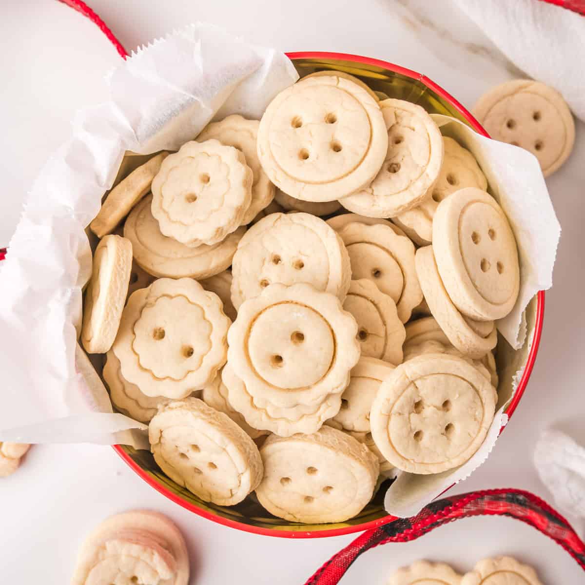 A round tin lined with white paper, filled with button-shaped sugar cookies, some with scalloped edges and holes to mimic button designs. A red ribbon is partially visible around the tin.