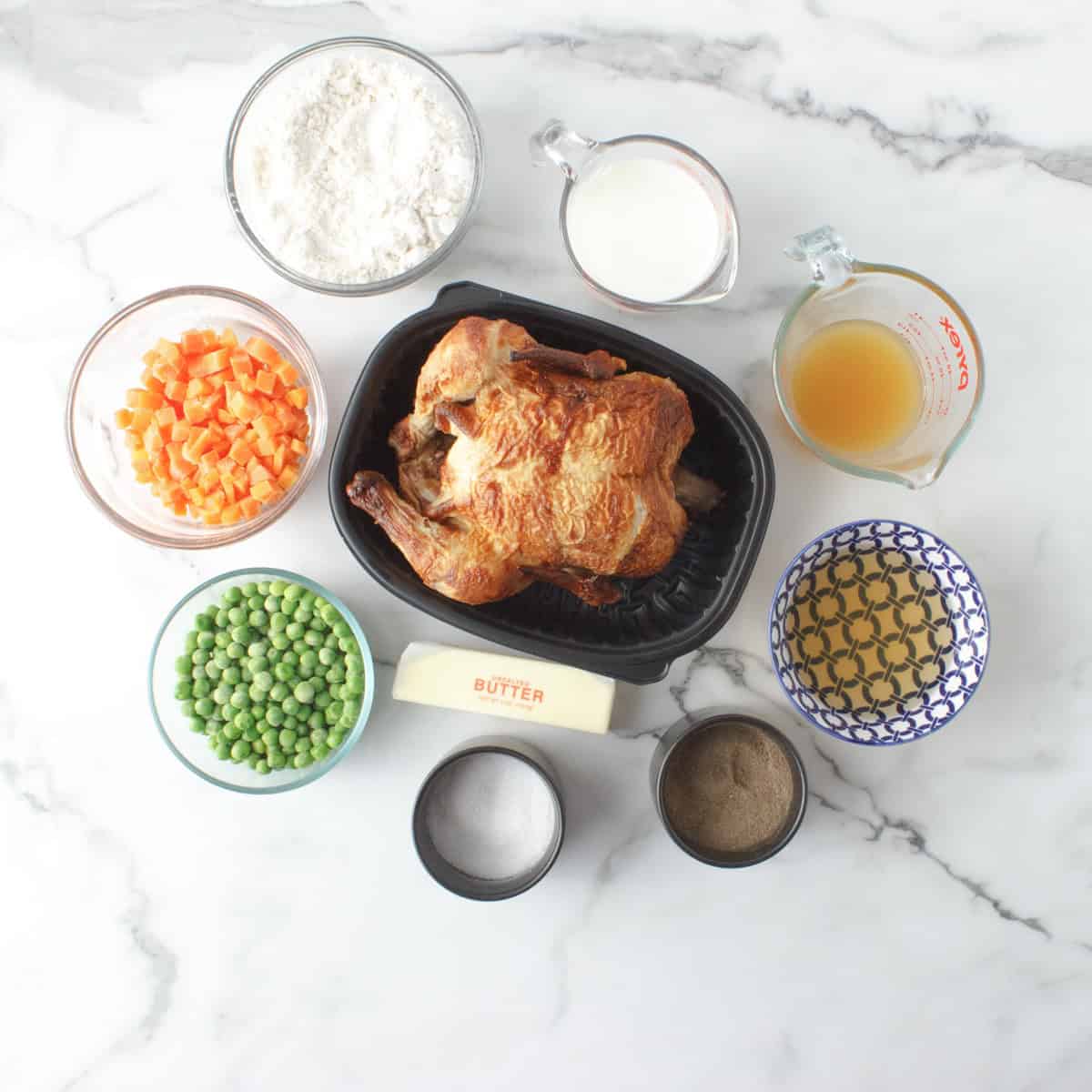 Overhead view of ingredients on a marble surface: whole roasted chicken, flour, milk, chicken broth, diced carrots, peas, butter, black pepper, salt, and a bowl of chopped onions.