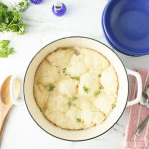 A white pot filled with fluffy dumplings garnished with herbs sits on a marble counter, surrounded by a blue bowl, utensils, parsley, and a salt shaker.