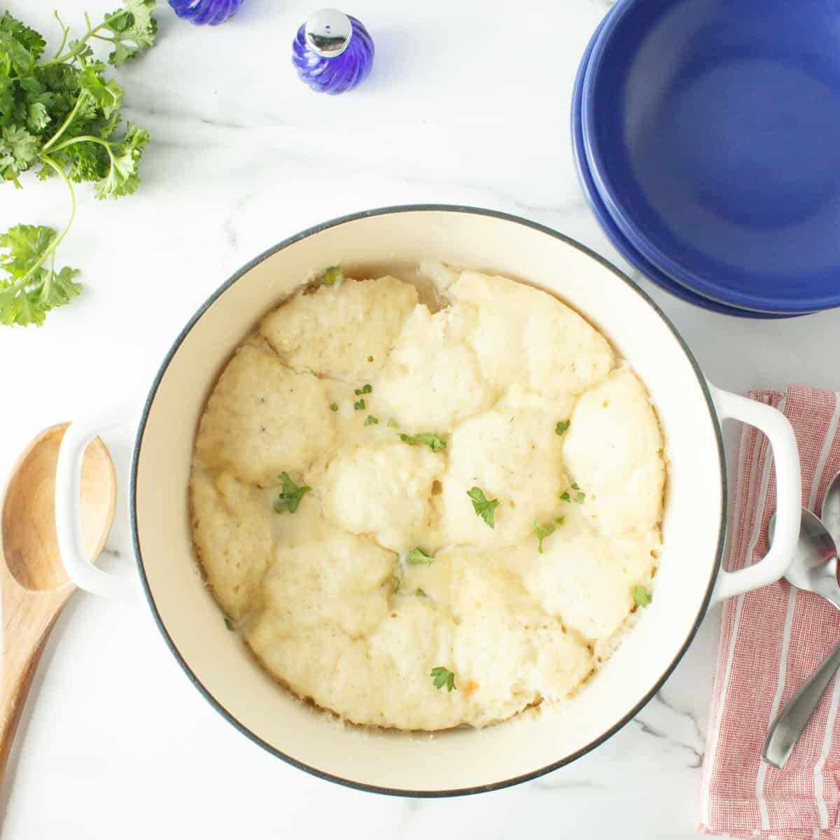 A white pot filled with fluffy dumplings garnished with herbs sits on a marble counter, surrounded by a blue bowl, utensils, parsley, and a salt shaker.