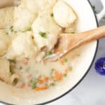 A white pot filled with creamy chicken and vegetable stew, topped with fluffy dumplings. A wooden spoon rests in the pot, revealing peas and carrots in the sauce.