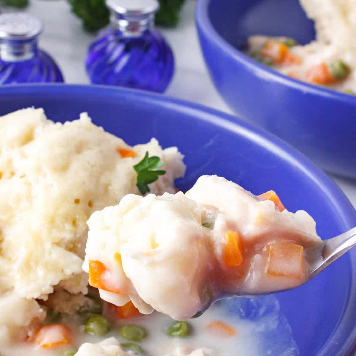 A spoonful of creamy chicken and dumplings, with carrots and peas, held above a blue bowl containing more of the dish. Two blue glass containers are in the background.