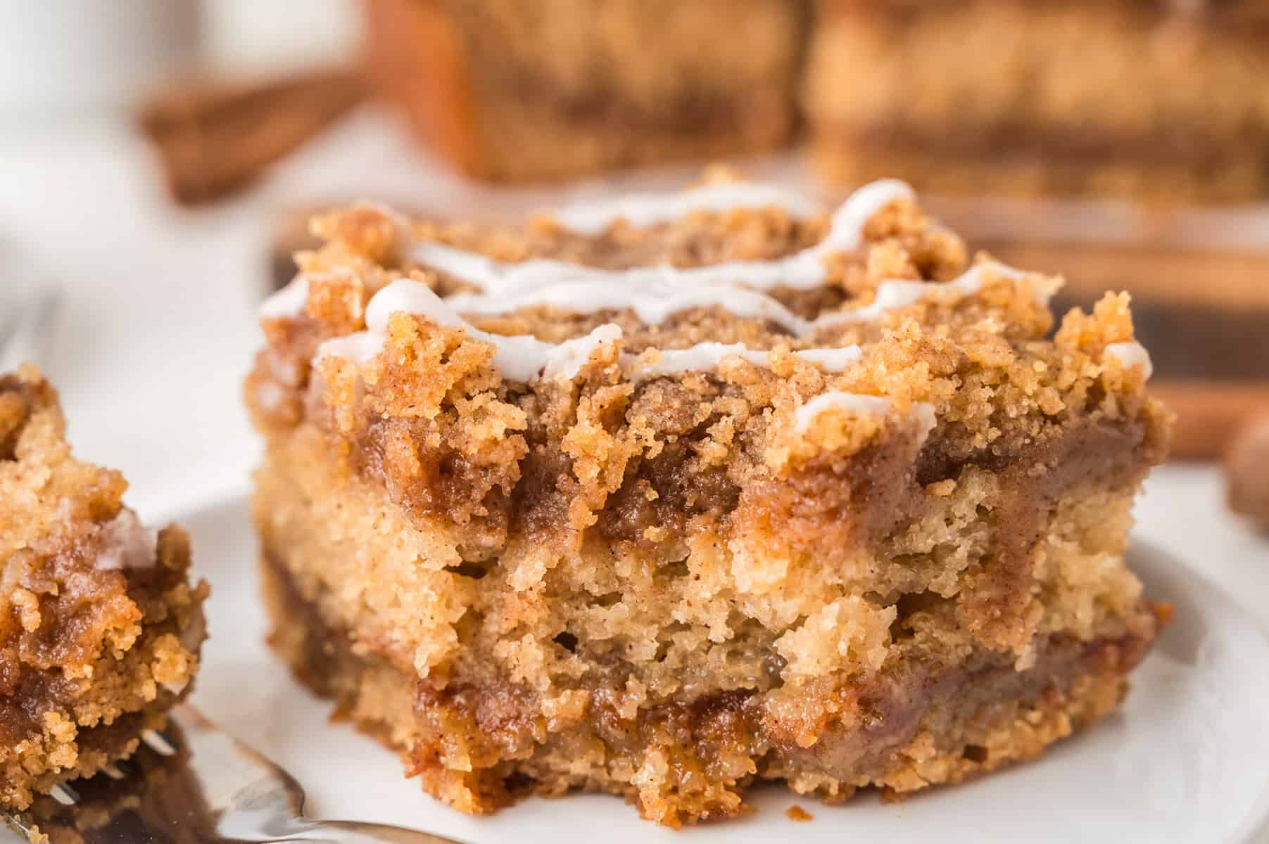 A close-up of a slice of cinnamon crumb cake with a drizzle of white icing on top, showing a soft, moist texture and layers of cinnamon filling and crumb topping.