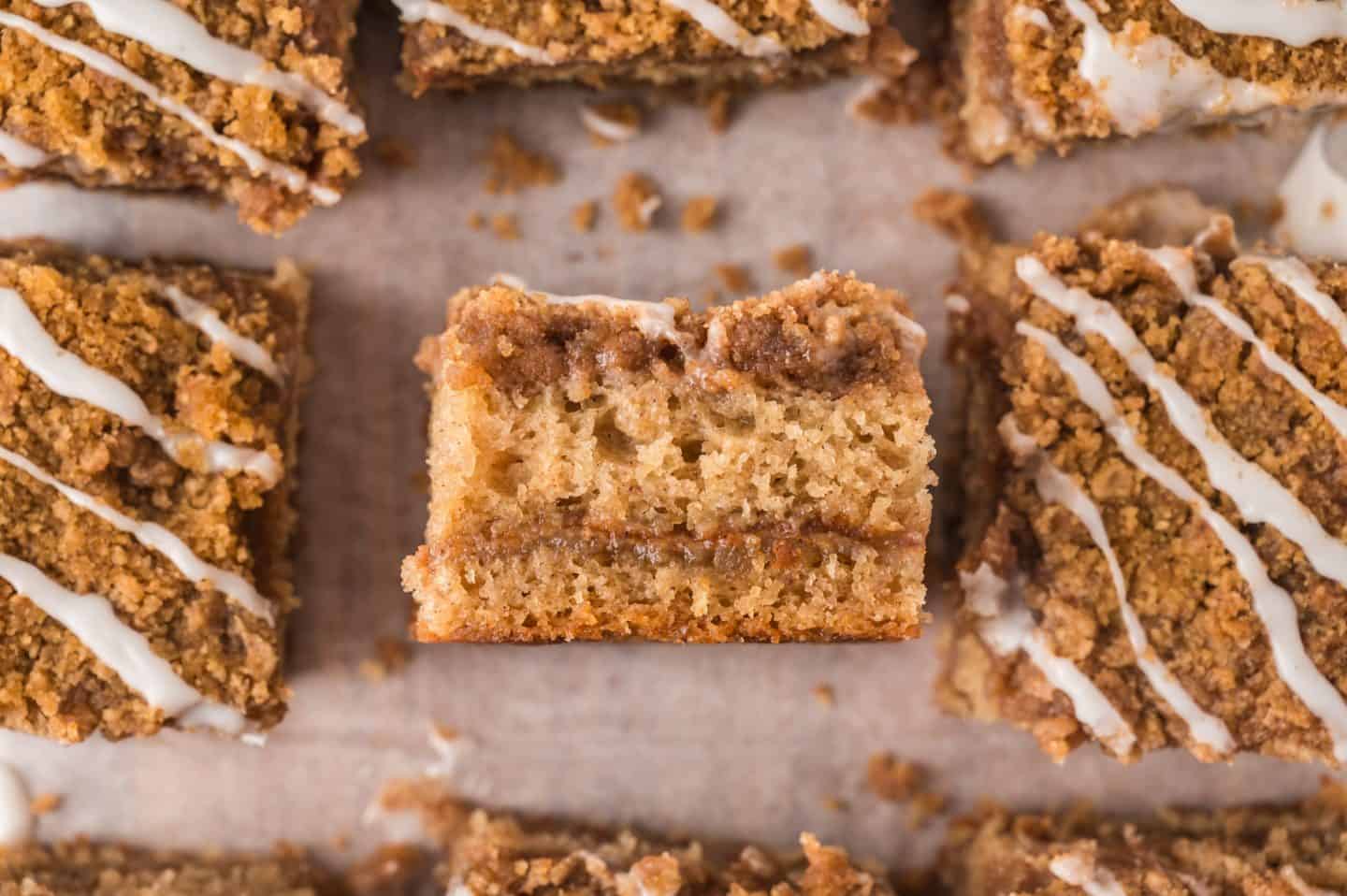 A close-up of sliced coffee cake squares with a crumbly streusel topping and white icing drizzles, displayed on parchment paper. One piece is turned to show the moist, layered interior.
