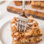 A fork is slicing into a piece of crumbly coffee cake with streusel topping and icing, set on a white plate. Another slice is visible in the background on a wooden board.