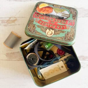 A vintage biscuit tin holds sewing items like buttons, a zipper, small scissors, thread, and a measuring tape. A spool of gray thread sits next to the tin on a light wooden surface.