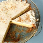 A glass pie dish with a creamy pie, topped with whipped cream and crumbs. One slice is cut and slightly removed from the whole pie, showing the layered filling and crust. The background is a light blue surface.