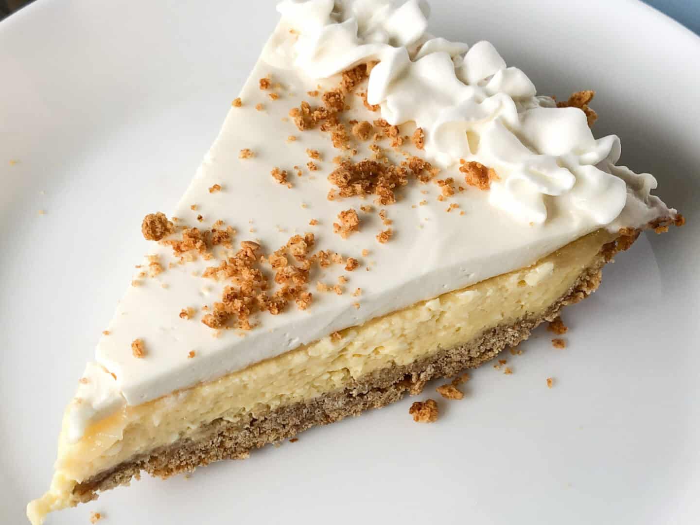 A slice of cheesecake with a graham cracker crust, creamy top layer, whipped cream, and sprinkled crumbs on a white plate.