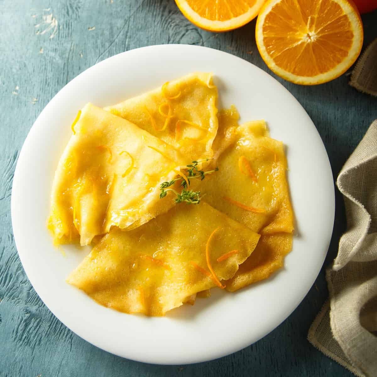 Crepes drizzled with orange glaze and garnished with zest and herbs on a white plate.