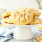 A pile of pastries topped with sliced almonds and white icing sit on a white cake stand. Blurred mugs, a blue-checked napkin, and more pastries are in the background on a light-colored table.