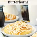A close-up of a homemade butterhorn pastry with icing and almond slices on a plate, surrounded by mugs and a syrup pitcher. Text: How to Make Homemade Butterhorns and theurbankitten.com.