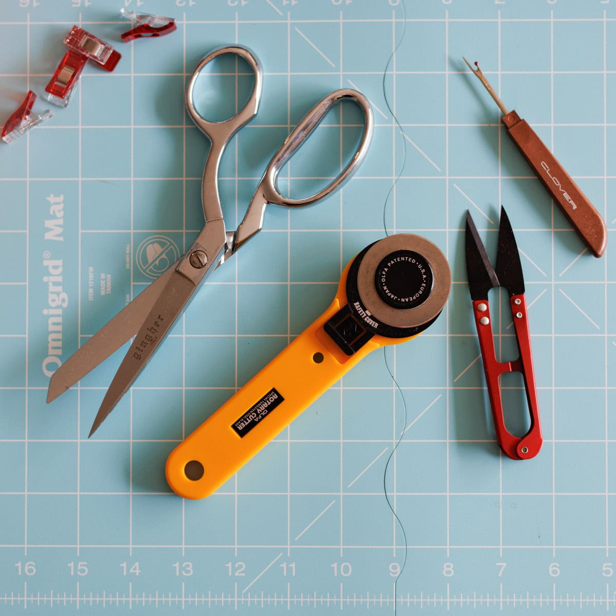 A pair of silver scissors, a yellow rotary cutter, red thread snips, a brown seam ripper, and red fabric clips are arranged on a blue Omnigrid cutting mat marked with a grid.
