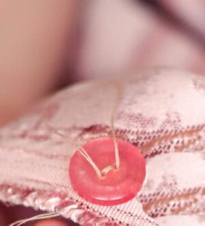 A close-up of a pink button being sewn onto pink fabric with a needle and yellow thread.