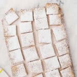 A batch of rectangular lemon bars dusted with powdered sugar, cut into squares and arranged on a marble surface next to a white and yellow striped towel.
