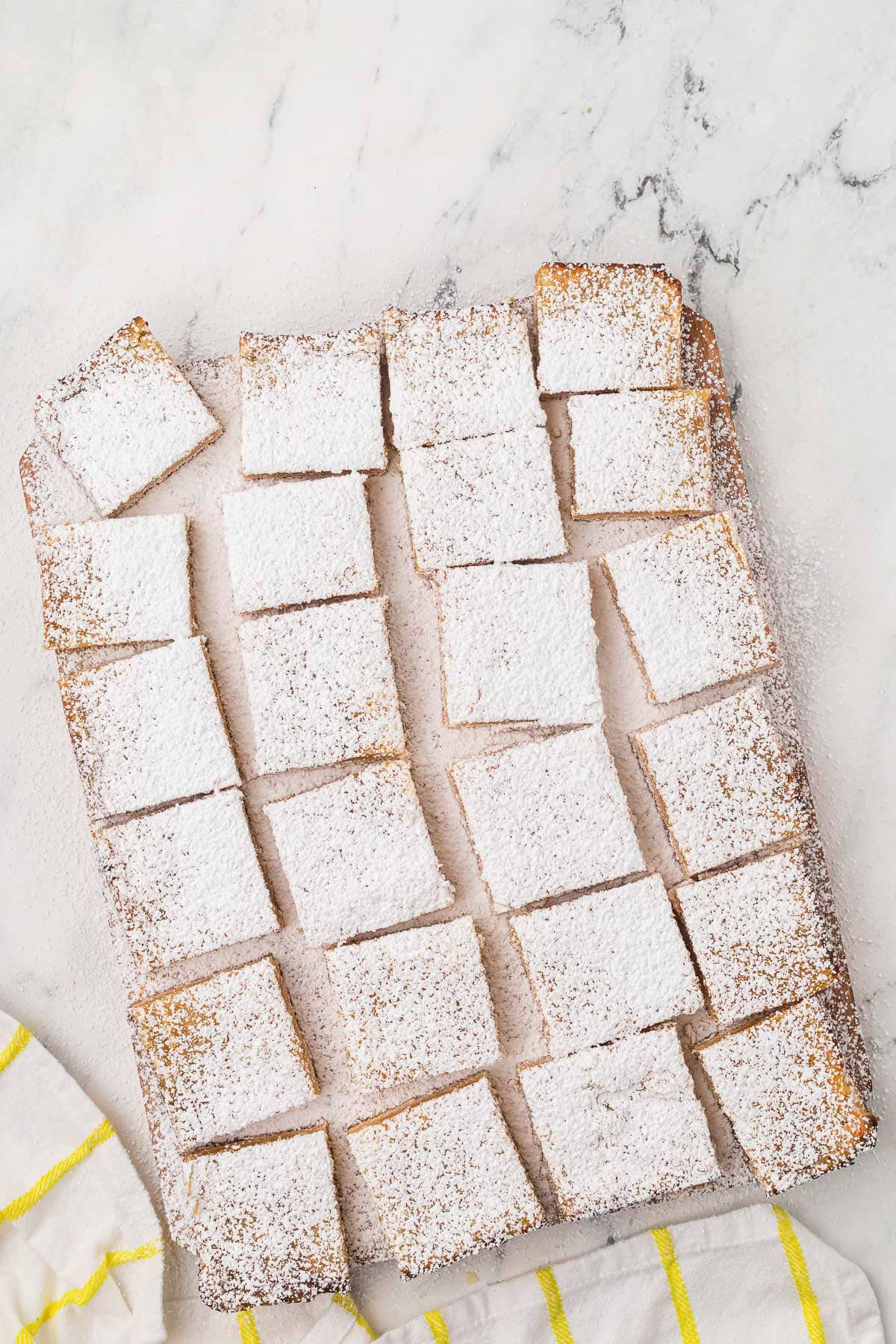 A batch of rectangular lemon bars dusted with powdered sugar, cut into squares and arranged on a marble surface next to a white and yellow striped towel.