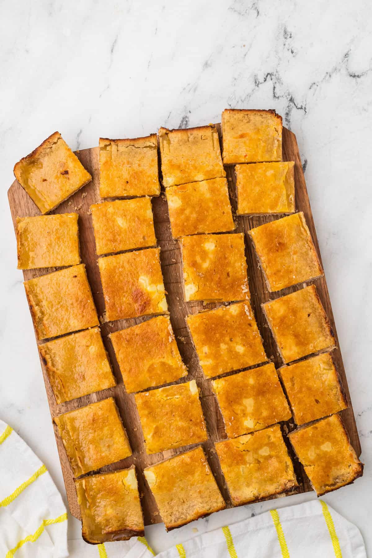 Twenty golden brown dessert bars are arranged in neat rows on a wooden board, with one bar slightly separated. The board sits on a marble surface beside a yellow-striped white cloth.