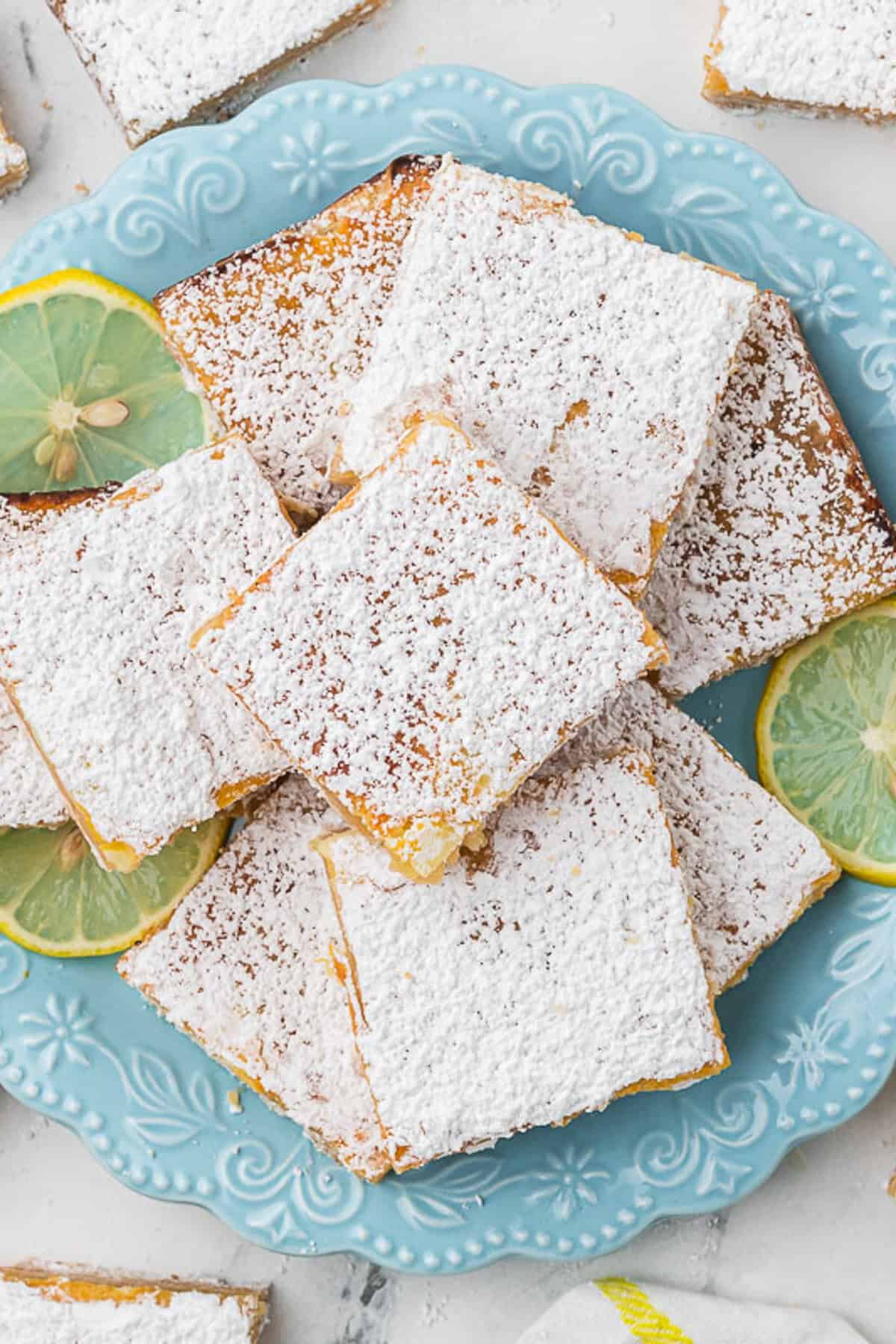 A blue plate holds several powdered sugar-dusted lemon bars, with lemon slices as garnish. More lemon bars are visible in the background on a light surface.