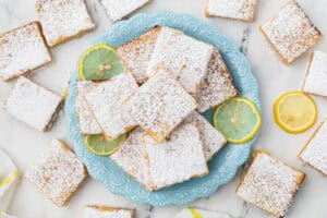 A blue plate holds powdered sugar-dusted lemon bars, surrounded by lemon slices. Extra lemon bars are scattered on a white marble surface.