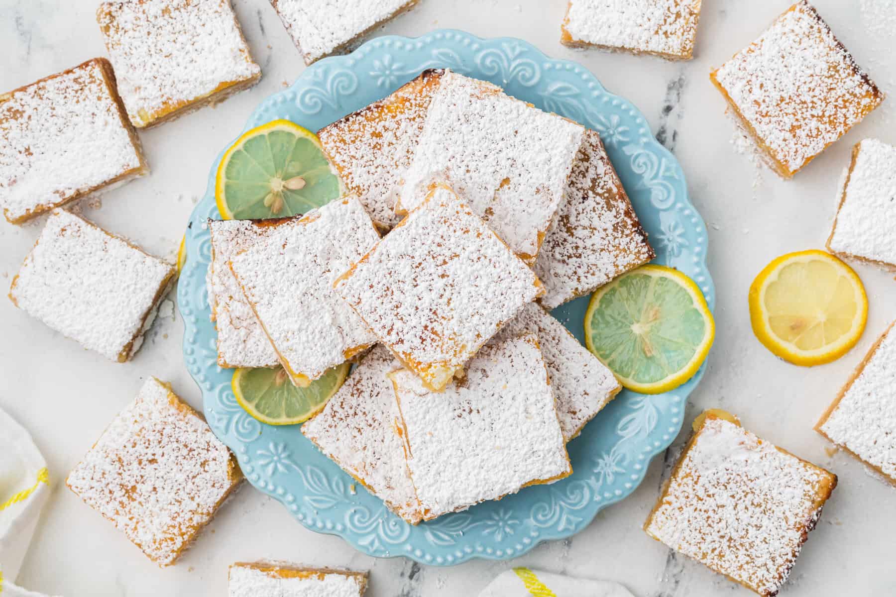 A blue plate holds powdered sugar-dusted lemon bars, surrounded by lemon slices. Extra lemon bars are scattered on a white marble surface.