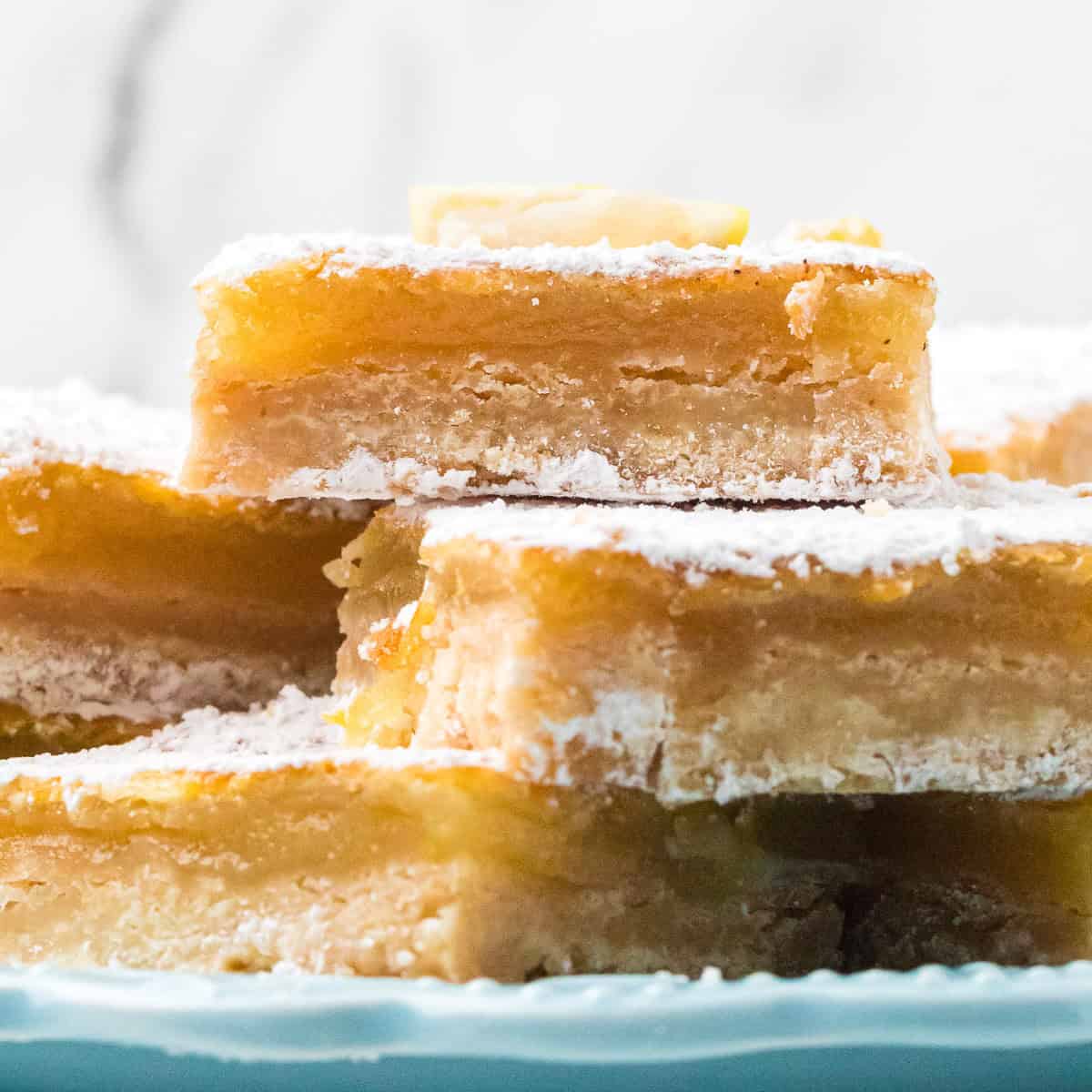 A close-up of stacked lemon bars topped with powdered sugar on a light blue plate, showing their golden, gooey layers and crumbly crust.