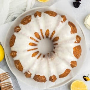 A round bundt cake with white icing drizzle sits on parchment paper. Lemon slices, a fork, and a dark flower are visible nearby on the light gray surface.