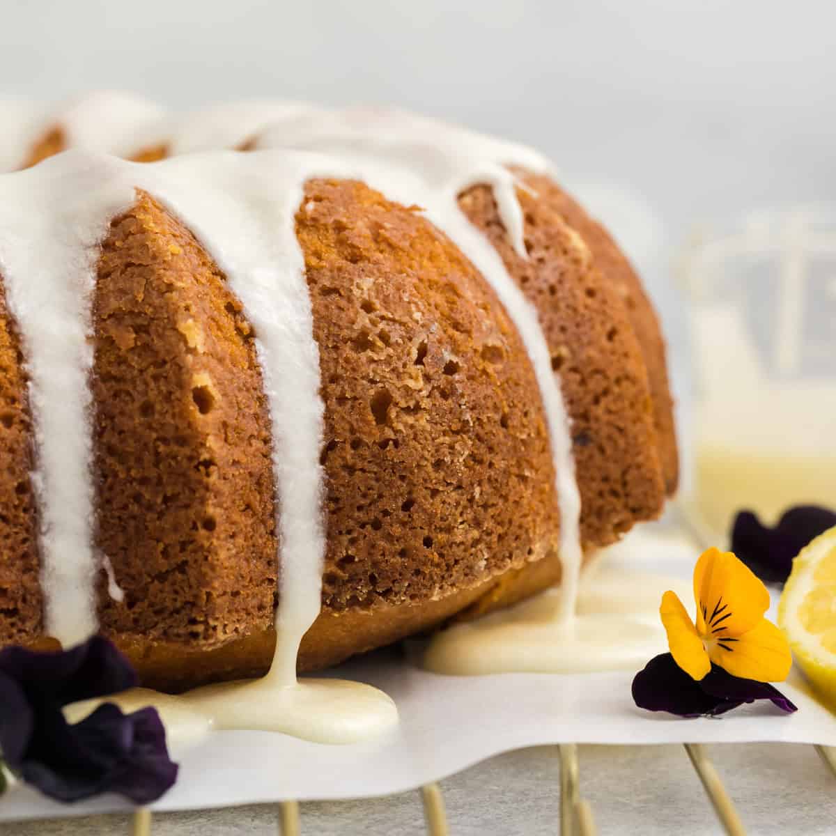 A close-up of a bundt cake with white icing drizzling down the sides, surrounded by edible flowers and a lemon slice on a wire rack.